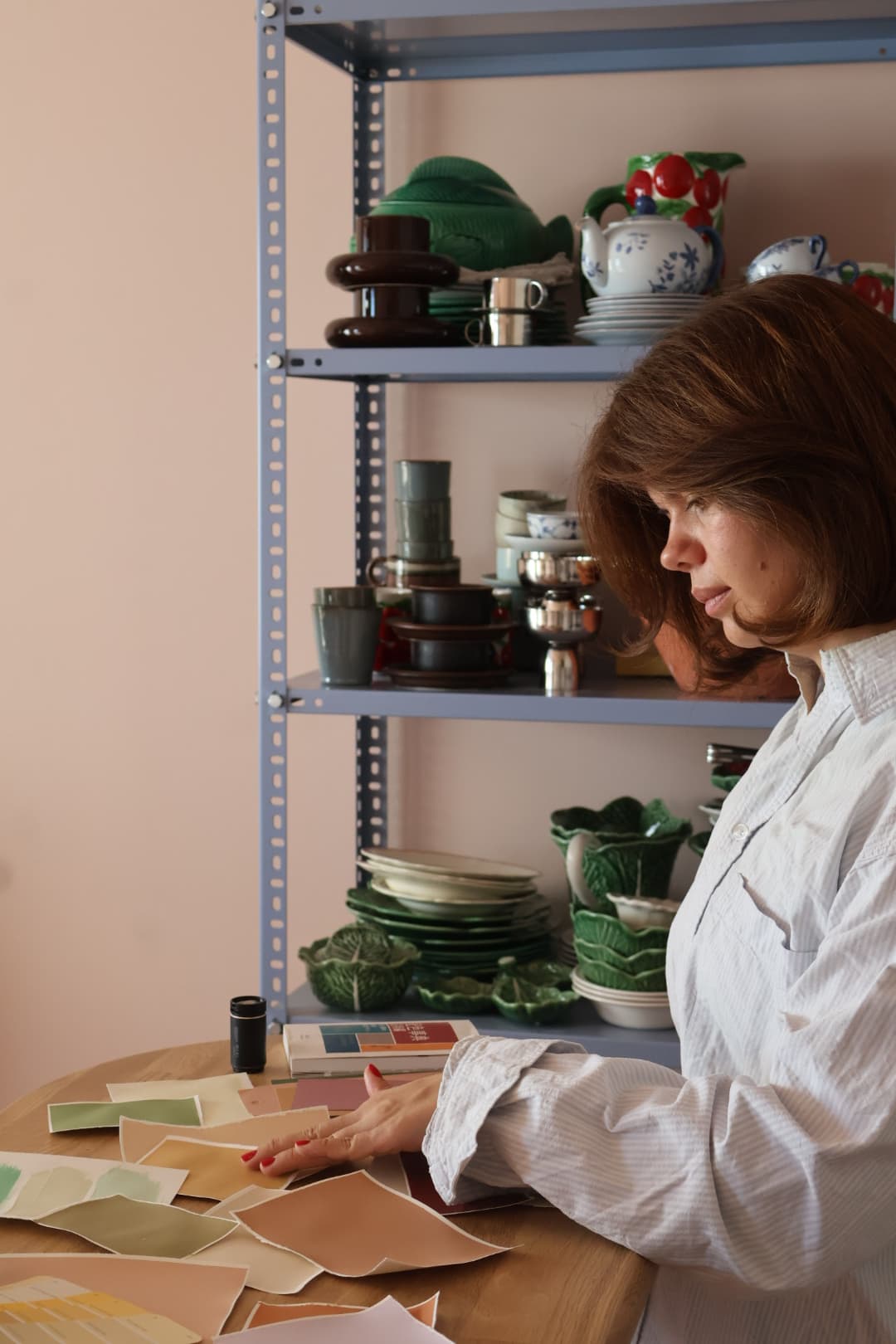 Sandra arbeider med fargeprøver ved et bord foran hyller med keramikk.